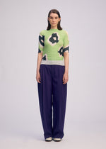 A woman with straight brown hair poses against a plain light background, wearing the ISSEY MIYAKE VERVE PLEATS TOP—featuring black and white abstract patterns—and loose dark purple pants with white shoes, ideal for everyday style.