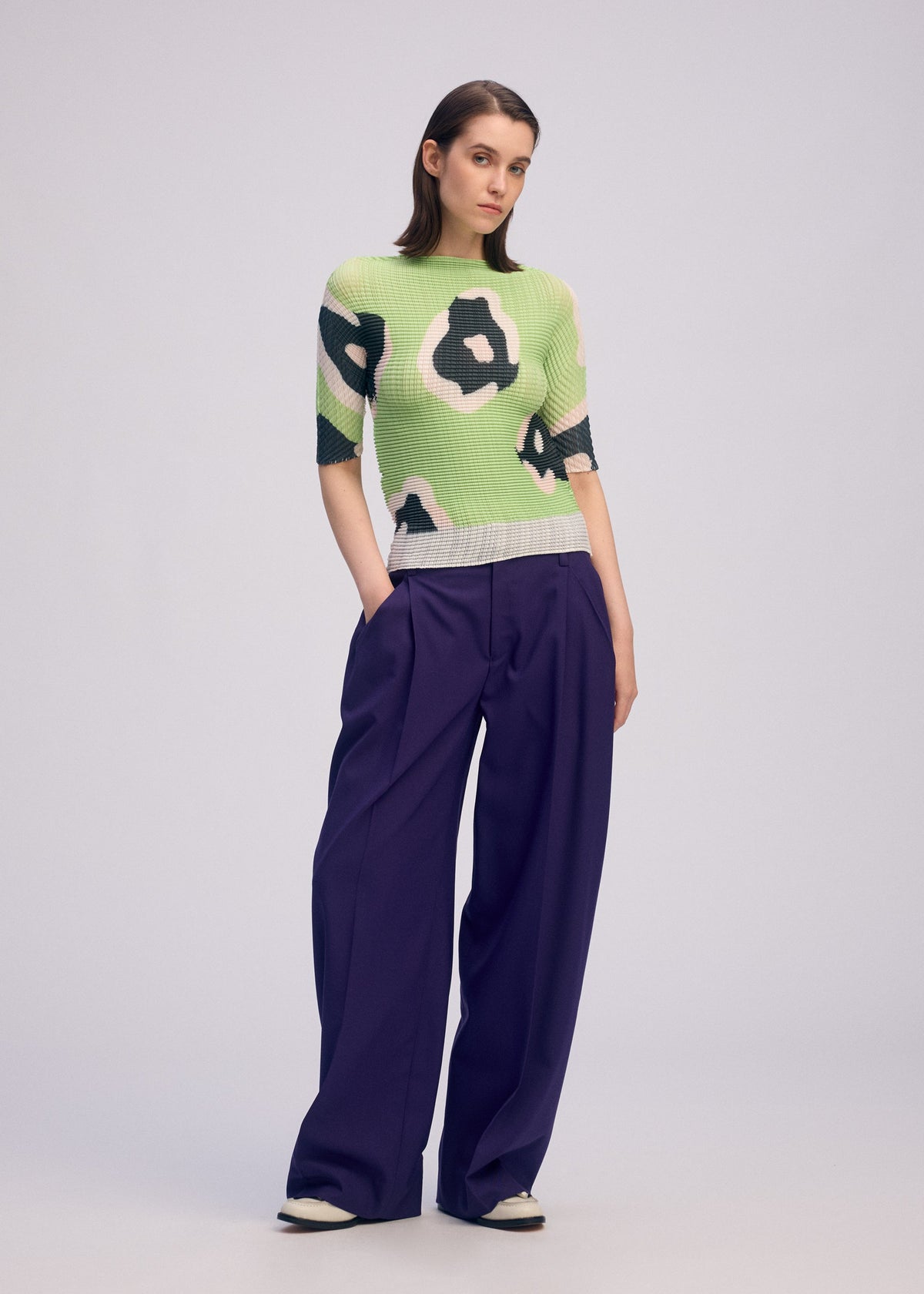 A woman stands against a plain background wearing the ISSEY MIYAKE VERVE PLEATS TOP with a textured green and white abstract pattern, styled with high-waisted, wide-leg purple trousers and white shoes for a chic everyday look. view 6 of 8