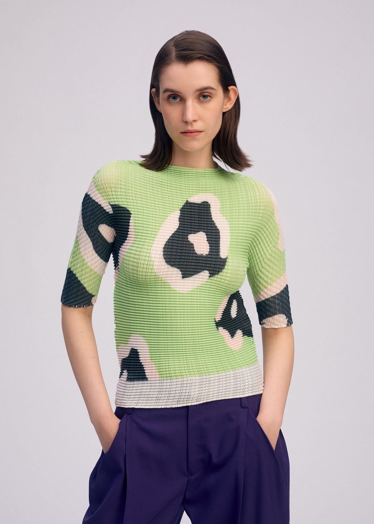 A woman with straight brown hair models the ISSEY MIYAKE VERVE PLEATS TOP, a light green pleated boat-neck top with bold black and white abstract patterns, styled with purple pants—an everyday statement against a light gray background. view 7 of 8
