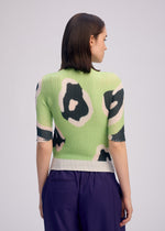 A person with straight brown hair is seen from the back, wearing ISSEY MIYAKE’s VERVE PLEATS TOP in a green and black floral print and purple pants, standing against a plain light background.