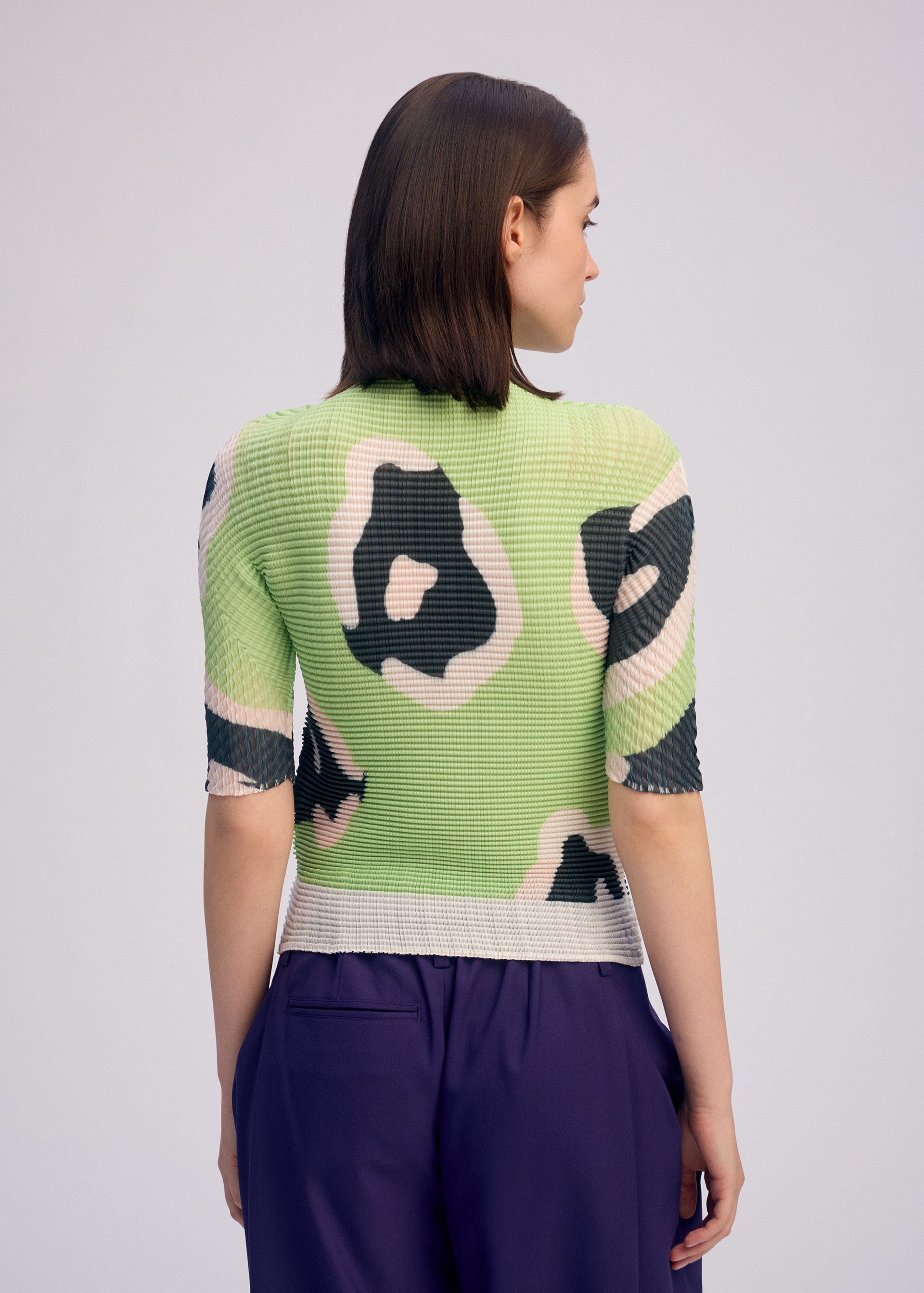 Zoomed view A person with straight brown hair is seen from the back, wearing ISSEY MIYAKE’s VERVE PLEATS TOP in a green and black floral print and purple pants, standing against a plain light background.