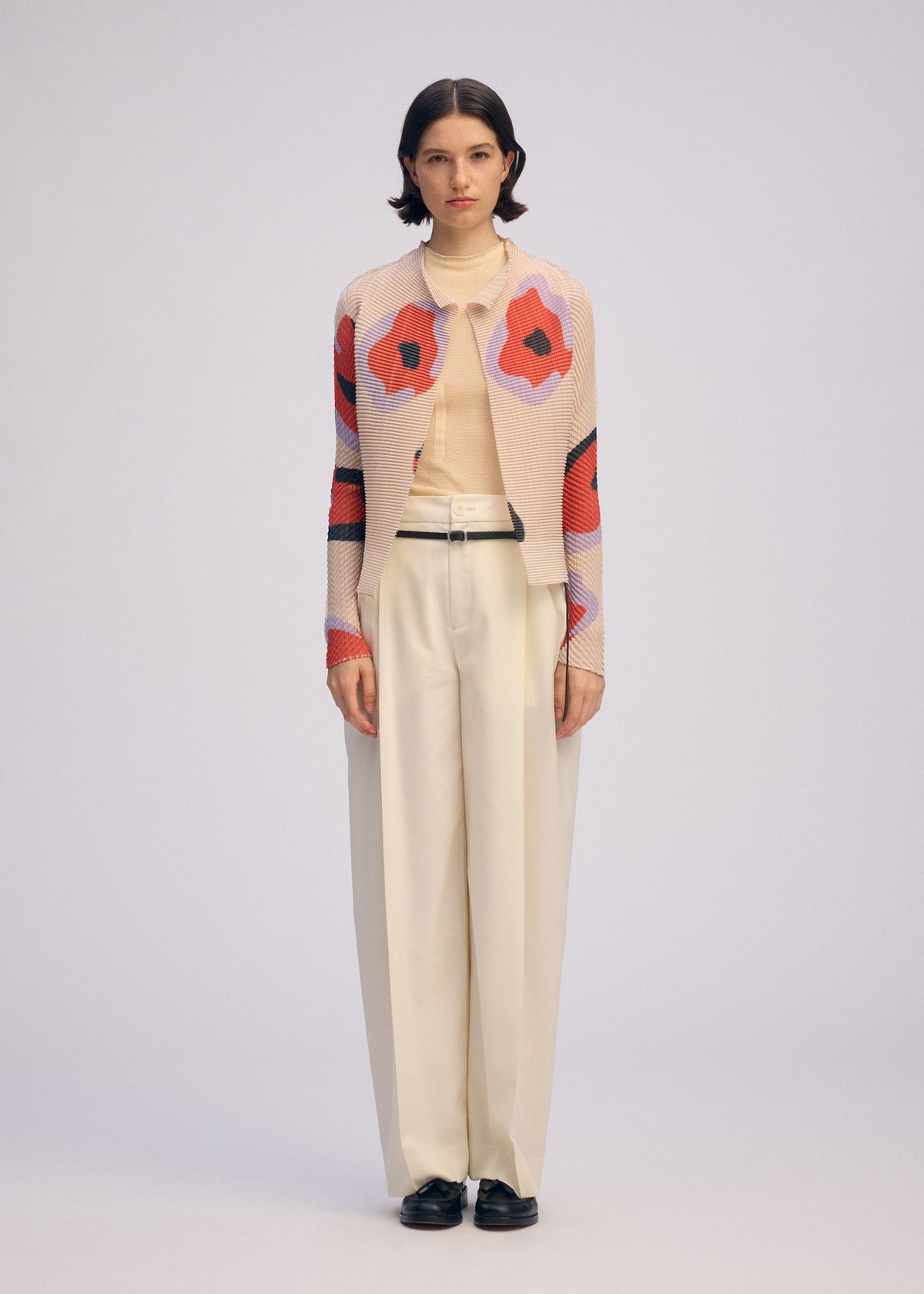A woman faces forward in a beige turtleneck, cream wide-leg trousers, black shoes, and the ISSEY MIYAKE VERVE PLEATS TOP—a pink pleated cardigan with bold red and black florals—against a plain light background. view 5 of 8
