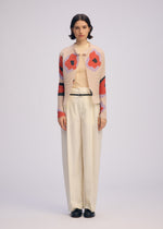 A woman faces forward in a beige turtleneck, cream wide-leg trousers, black shoes, and the ISSEY MIYAKE VERVE PLEATS TOP—a pink pleated cardigan with bold red and black florals—against a plain light background.