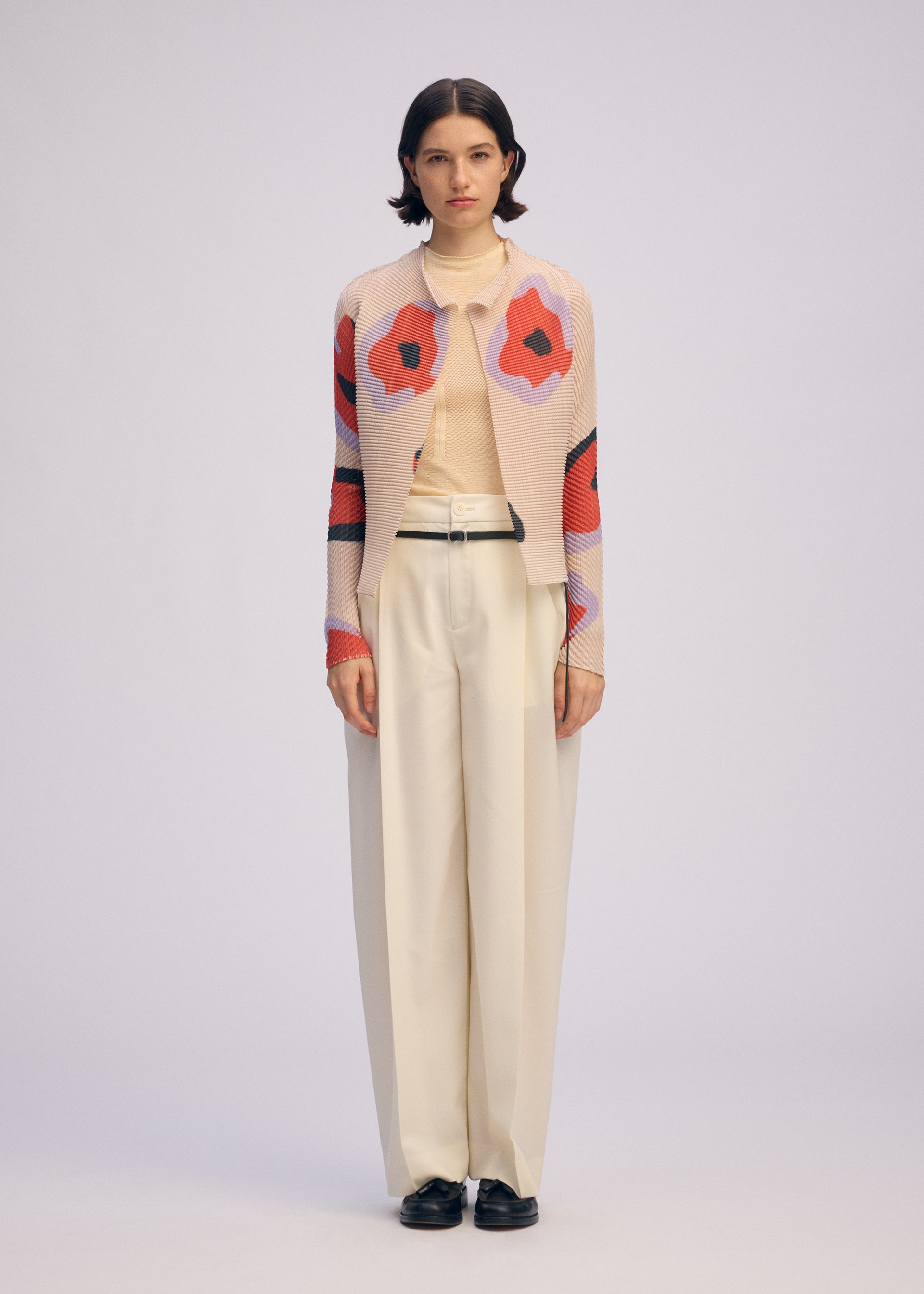 Zoomed view A woman faces forward in a beige turtleneck, cream wide-leg trousers, black shoes, and the ISSEY MIYAKE VERVE PLEATS TOP—a pink pleated cardigan with bold red and black florals—against a plain light background.