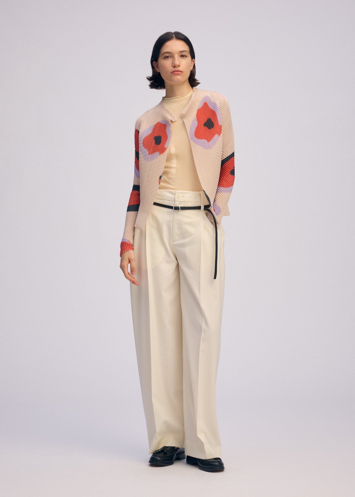 A person stands against a plain background wearing the ISSEY MIYAKE VERVE PLEATS TOP—a beige, red, and black floral pleated cardigan—paired with a beige turtleneck, wide-leg cream trousers, and black shoes. view 6 of 8