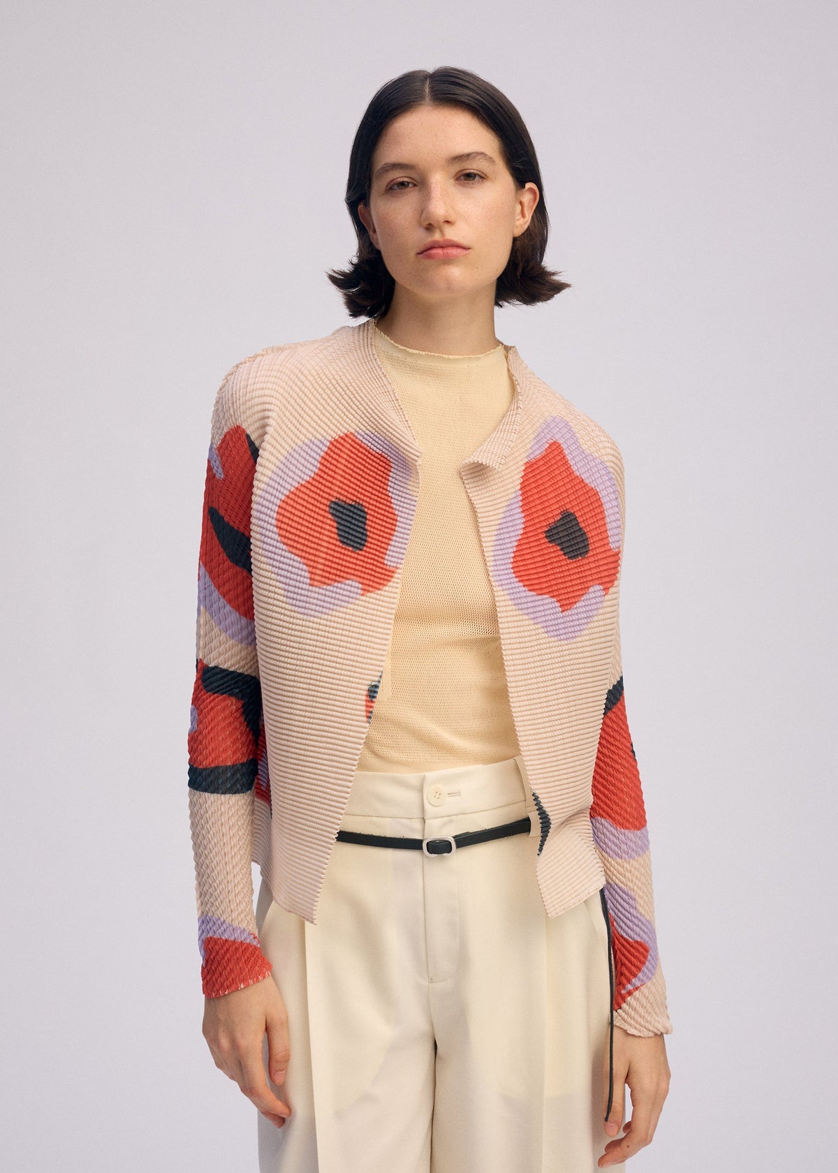 A woman with short dark hair models the VERVE PLEATS TOP by ISSEY MIYAKE—a beige turtleneck paired with cream pants, a black belt, and a floral print cardigan with bold red and black patterns—standing against a plain background. view 7 of 8