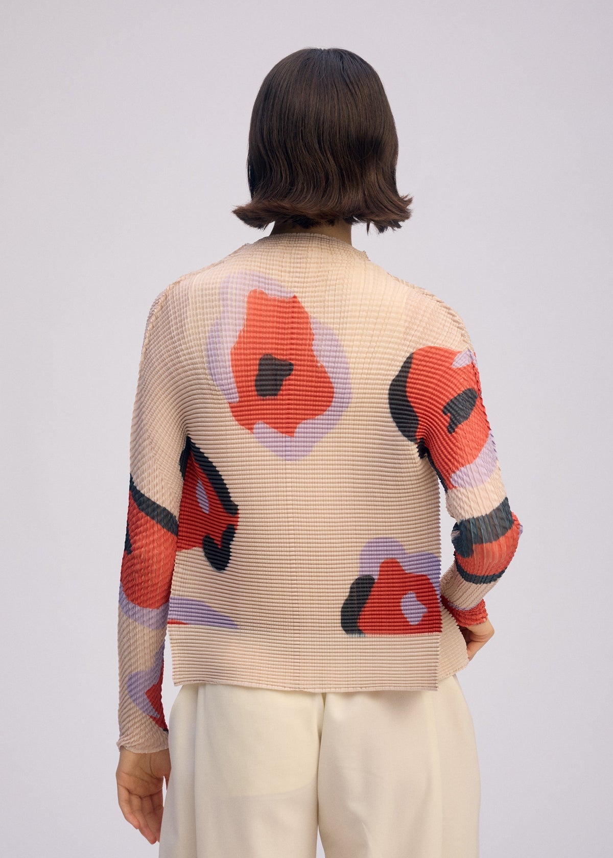A person with short, dark hair is seen from the back wearing the ISSEY MIYAKE VERVE PLEATS TOP, a beige pleated cardigan with bold red, black, and purple abstract floral prints, paired with loose white pants. view 8 of 8