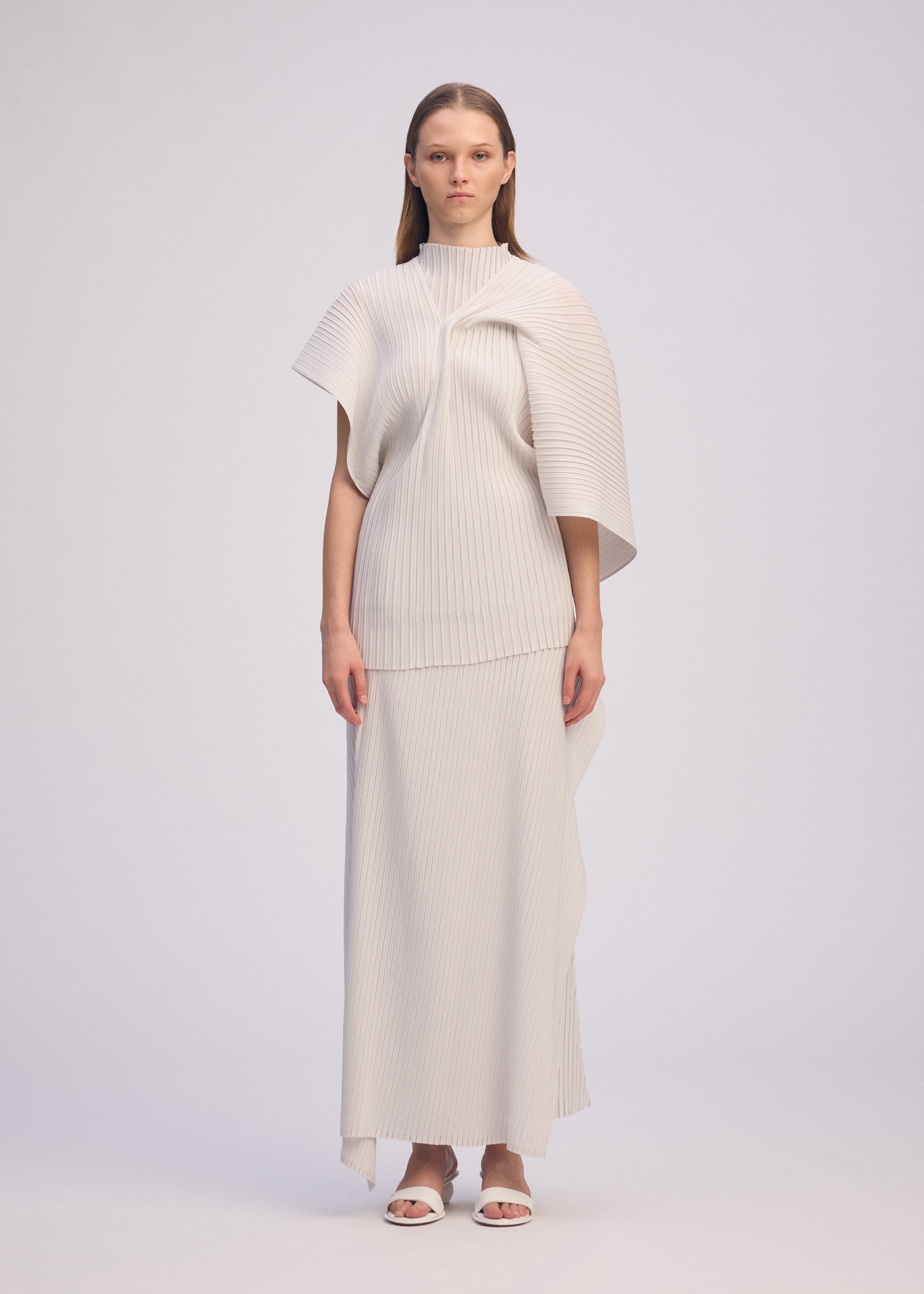 Zoomed view A woman stands against a plain background wearing ISSEY MIYAKE’s PLEATED ON PLEATS ribbed, high-neck top with a cape-like sleeve and a matching long skirt. She has straight hair and white sandals.