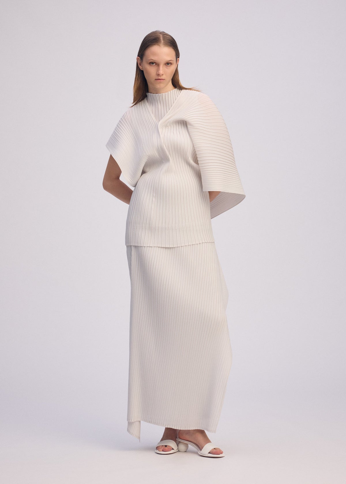 A woman with straight, long hair stands against a plain background wearing the ISSEY MIYAKE PLEATED ON PLEATS ivory ensemble featuring a structured, pleated top with asymmetrical sleeves and a matching long skirt, paired with white sandals. view 6 of 10