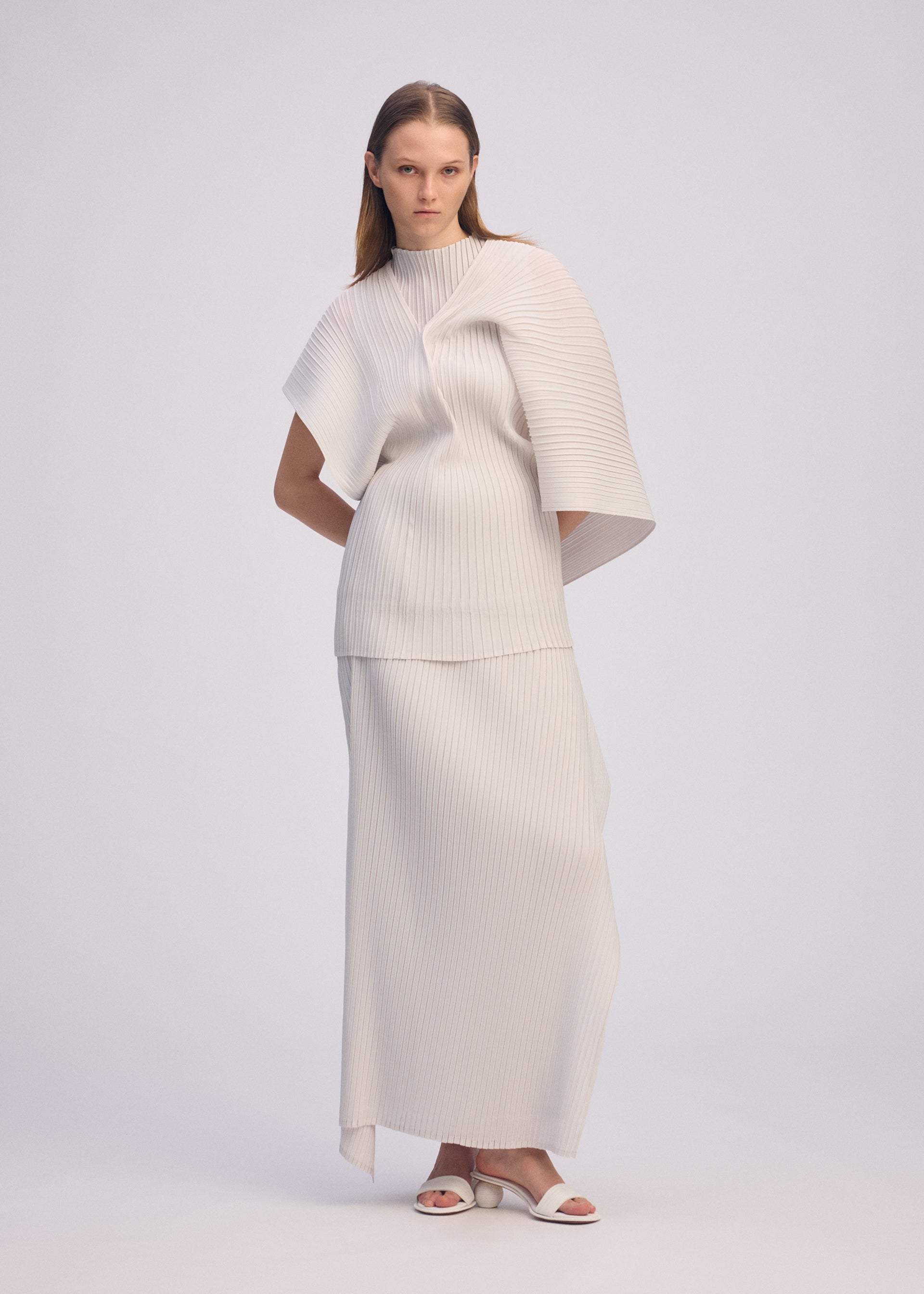 Zoomed view A woman with straight, long hair stands against a plain background wearing the ISSEY MIYAKE PLEATED ON PLEATS ivory ensemble featuring a structured, pleated top with asymmetrical sleeves and a matching long skirt, paired with white sandals.