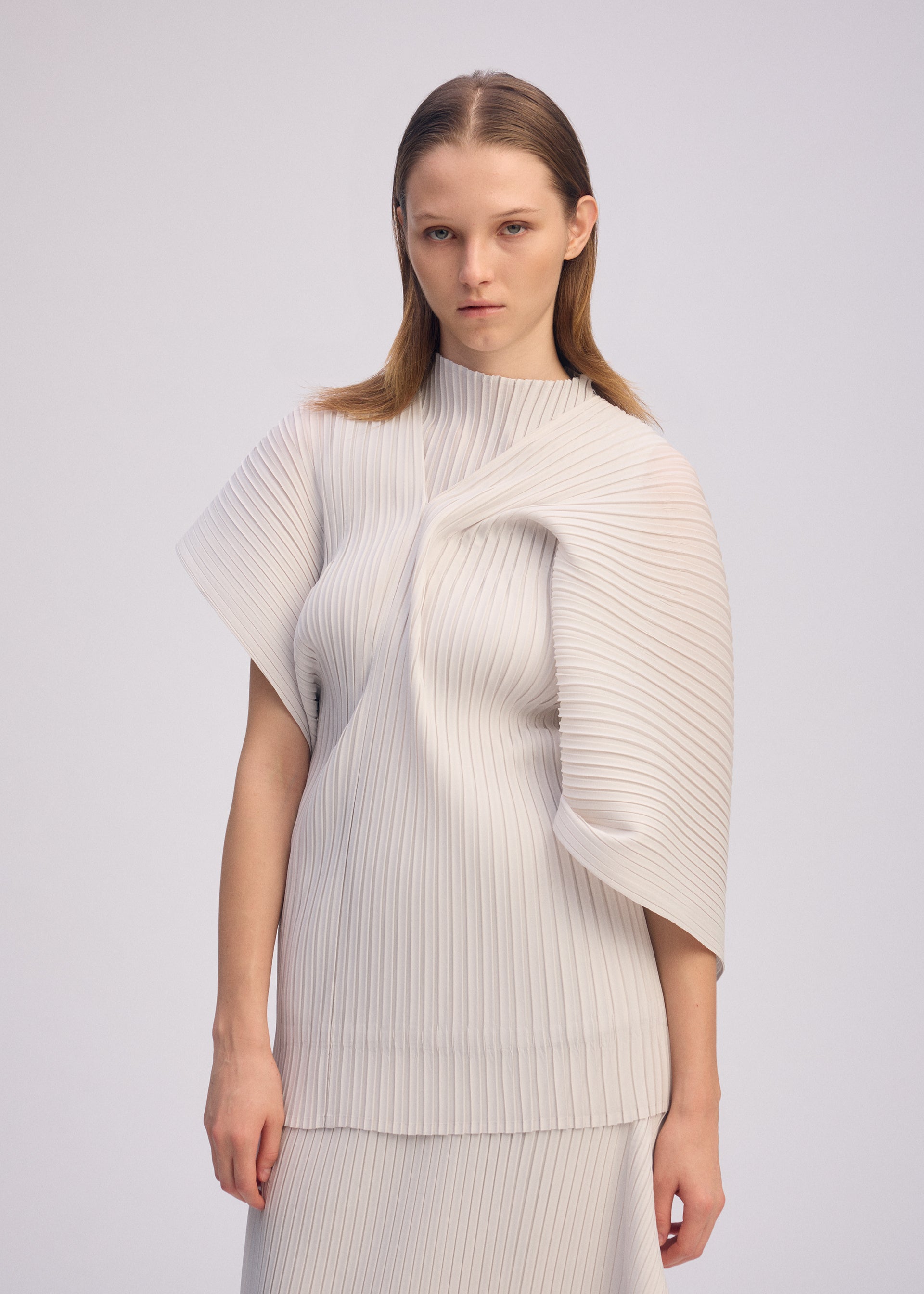 Zoomed view A woman with straight, light brown hair wears ISSEY MIYAKE’s PLEATED ON PLEATS set—an asymmetrical white top and matching skirt—in front of a plain, light background. She has a neutral expression.