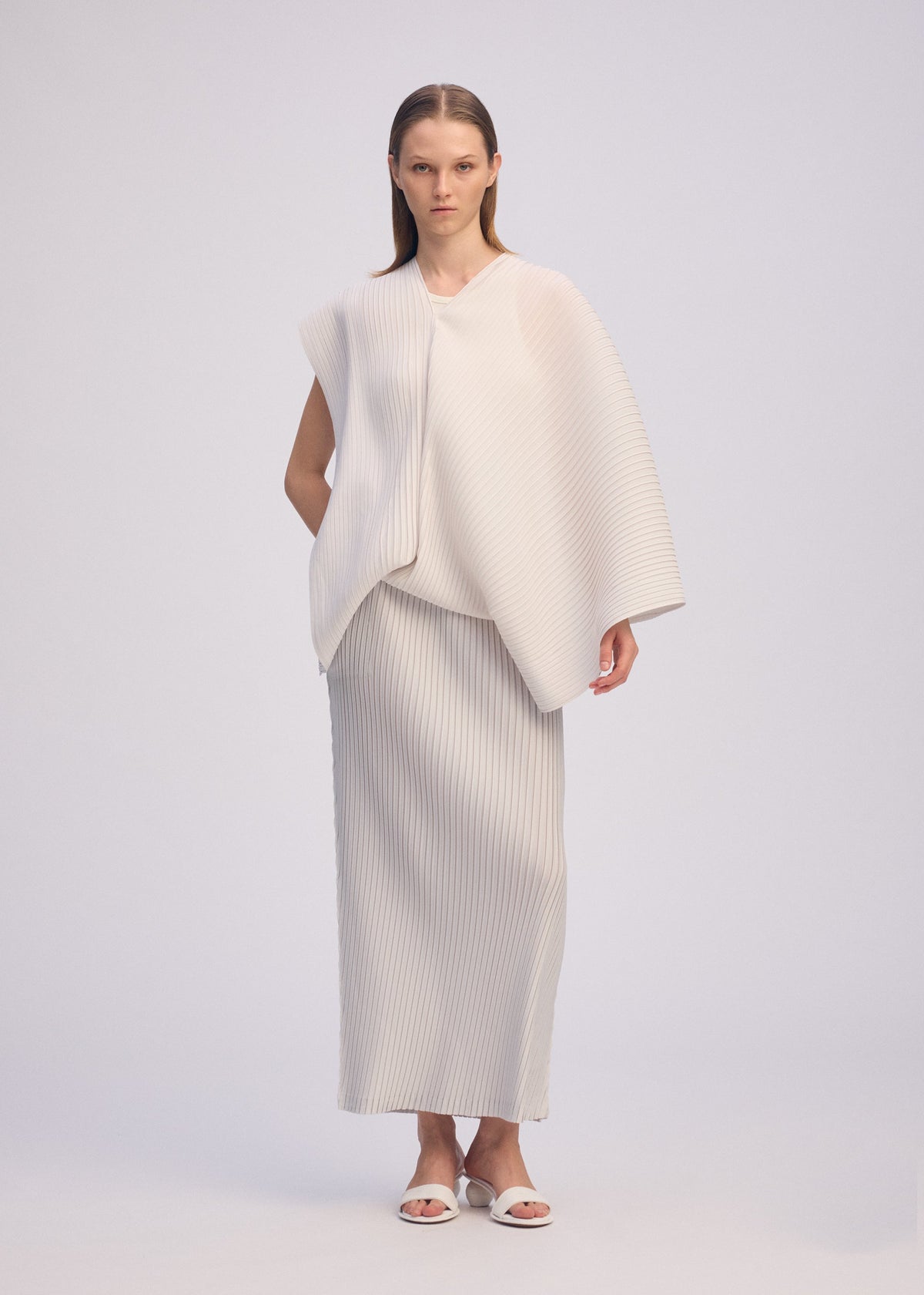 A woman stands facing forward, wearing ISSEY MIYAKE's PLEATED ON PLEATS ensemble: a loose, off-white asymmetrical pleated top and matching long skirt. She has straight hair, white sandals, and is posed against a plain light background. view 9 of 10