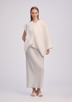 A woman stands facing forward, wearing ISSEY MIYAKE's PLEATED ON PLEATS ensemble: a loose, off-white asymmetrical pleated top and matching long skirt. She has straight hair, white sandals, and is posed against a plain light background.