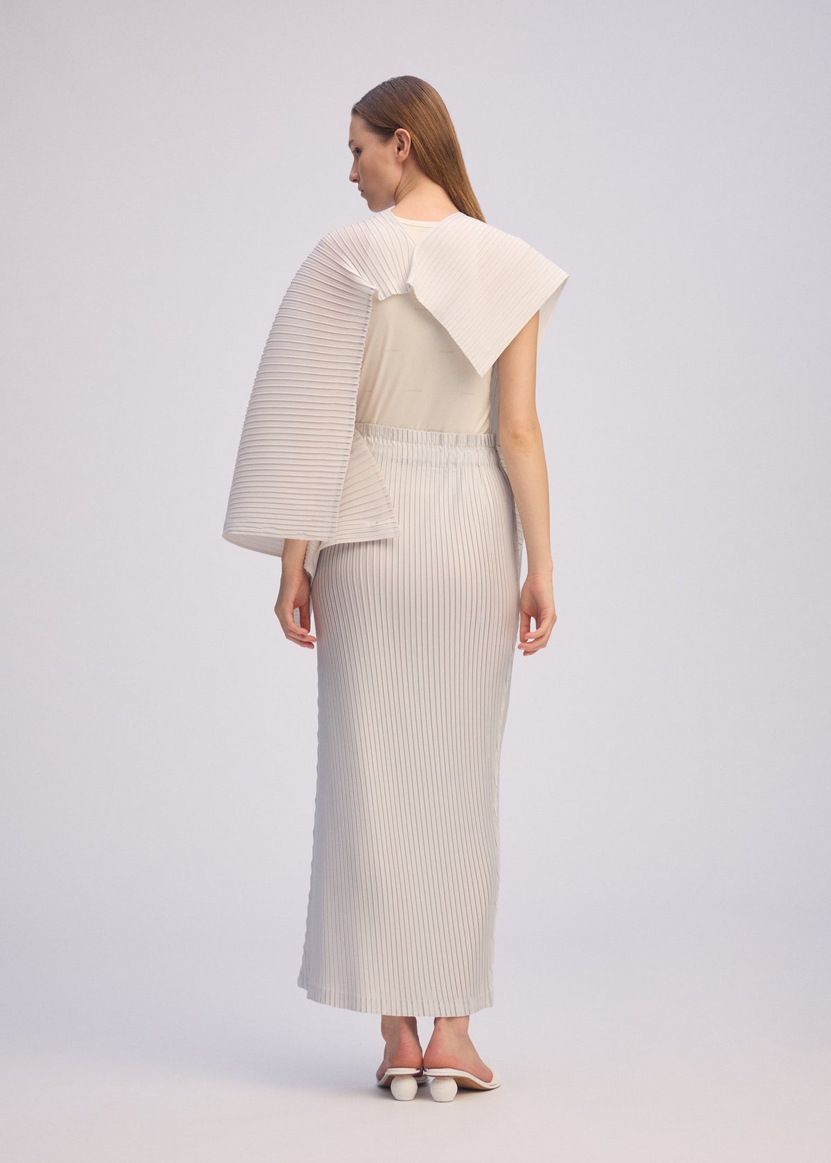 A woman faces away, wearing ISSEY MIYAKE’s PLEATED ON PLEATS set: a textured off-white high-neck top with sculptural pleated sleeves and a long pleated skirt. She pairs it with white heeled sandals against a plain background. view 10 of 10