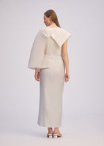 A woman faces away, wearing ISSEY MIYAKE’s PLEATED ON PLEATS set: a textured off-white high-neck top with sculptural pleated sleeves and a long pleated skirt. She pairs it with white heeled sandals against a plain background.