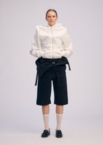 A person poses against a plain background, wearing ISSEY MIYAKE's GENERIC WEAR hoodie with unique details, paired with black knee-length shorts, white knee-high socks, and black loafers; hands in pockets, expression neutral.