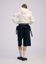 A person faces away wearing ISSEY MIYAKE’s GENERIC WEAR hoodie, black knee-length shorts, a black jacket tied at the waist, sheer white socks, and black loafers against a plain light backdrop with unique detailing.