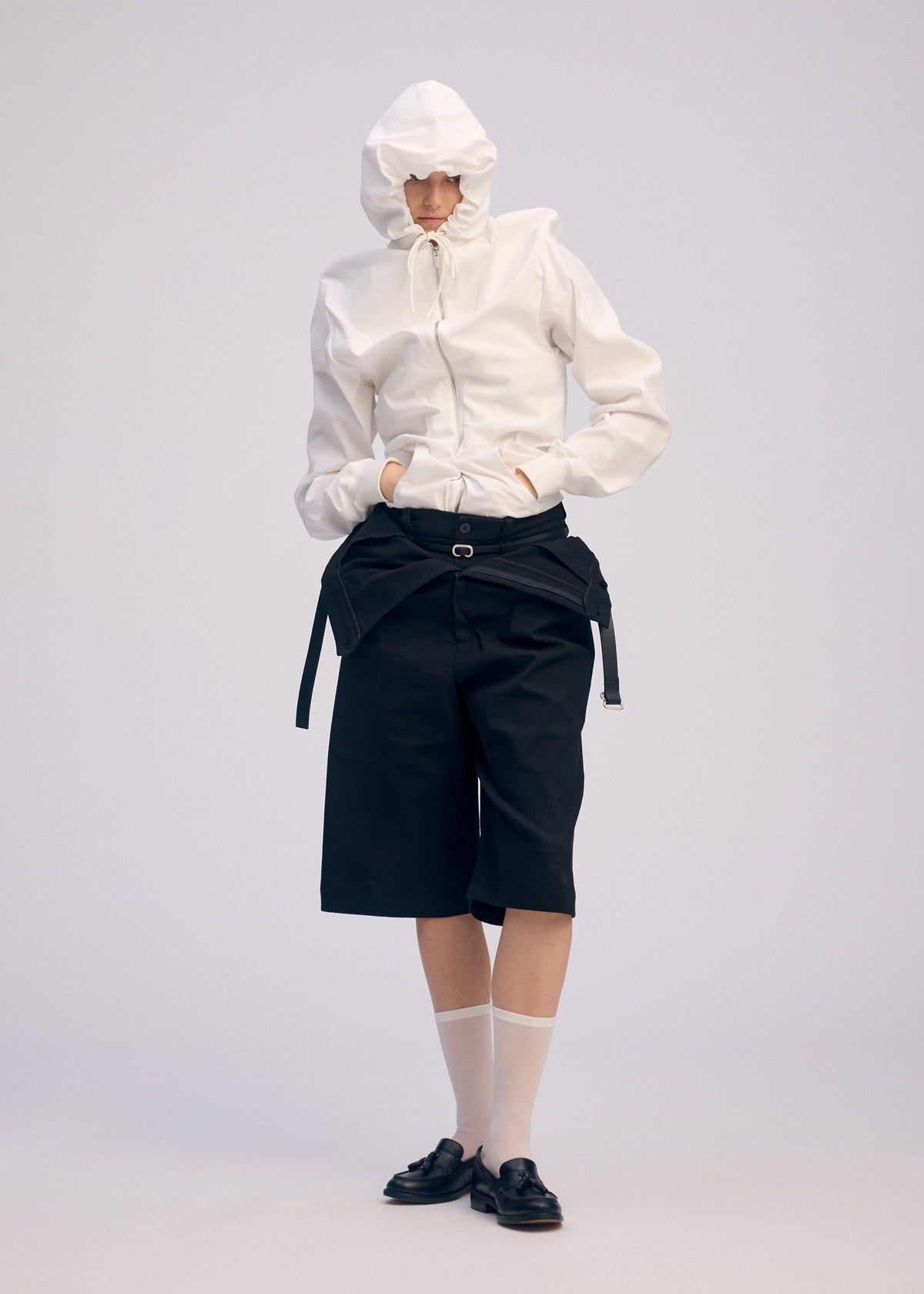 A person poses against a plain background in ISSEY MIYAKE's GENERIC WEAR: a playful hoodie with a white hood, black knee-length shorts, sheer white socks, and black loafers, hands tucked in pockets. view 8 of 10