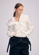 A person with light brown hair in a ponytail wears ISSEY MIYAKE’s GENERIC WEAR: a white zip-up jacket with unique detailing, partially unzipped off one shoulder, and black belted pants, standing against a plain light background.