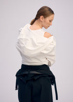 A person with light brown hair in a low ponytail models ISSEY MIYAKE's GENERIC WEAR: a white, one-shoulder top and black pants, with a playful hoodie tied at the waist, looking over their shoulder.