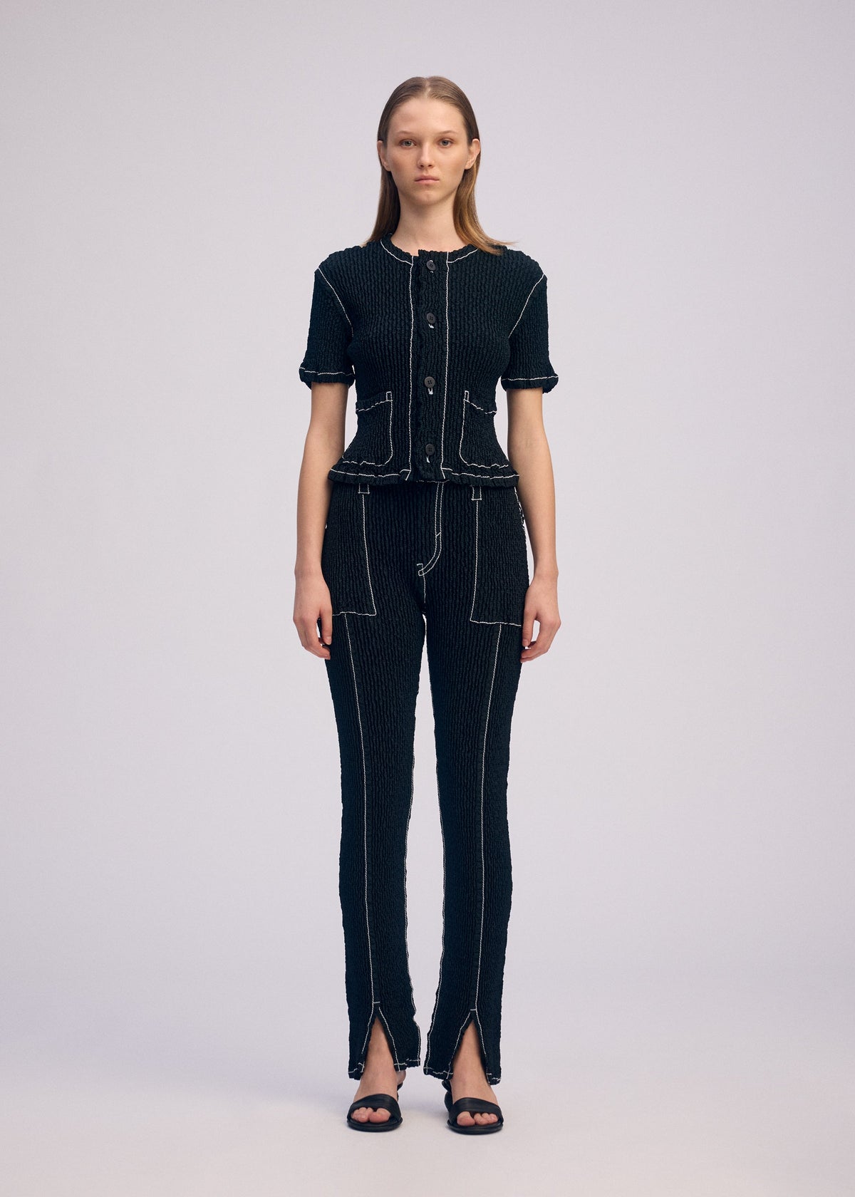 A woman with straight hair wears the ISSEY MIYAKE WAVELET ENSEMBLE—a jacquard stretch short-sleeved button-up top and matching pants with white stitching, front pockets, pant hem slits—and black sandals. view 5 of 7