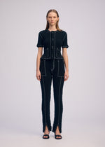 A woman with straight hair wears the ISSEY MIYAKE WAVELET ENSEMBLE—a jacquard stretch short-sleeved button-up top and matching pants with white stitching, front pockets, pant hem slits—and black sandals.