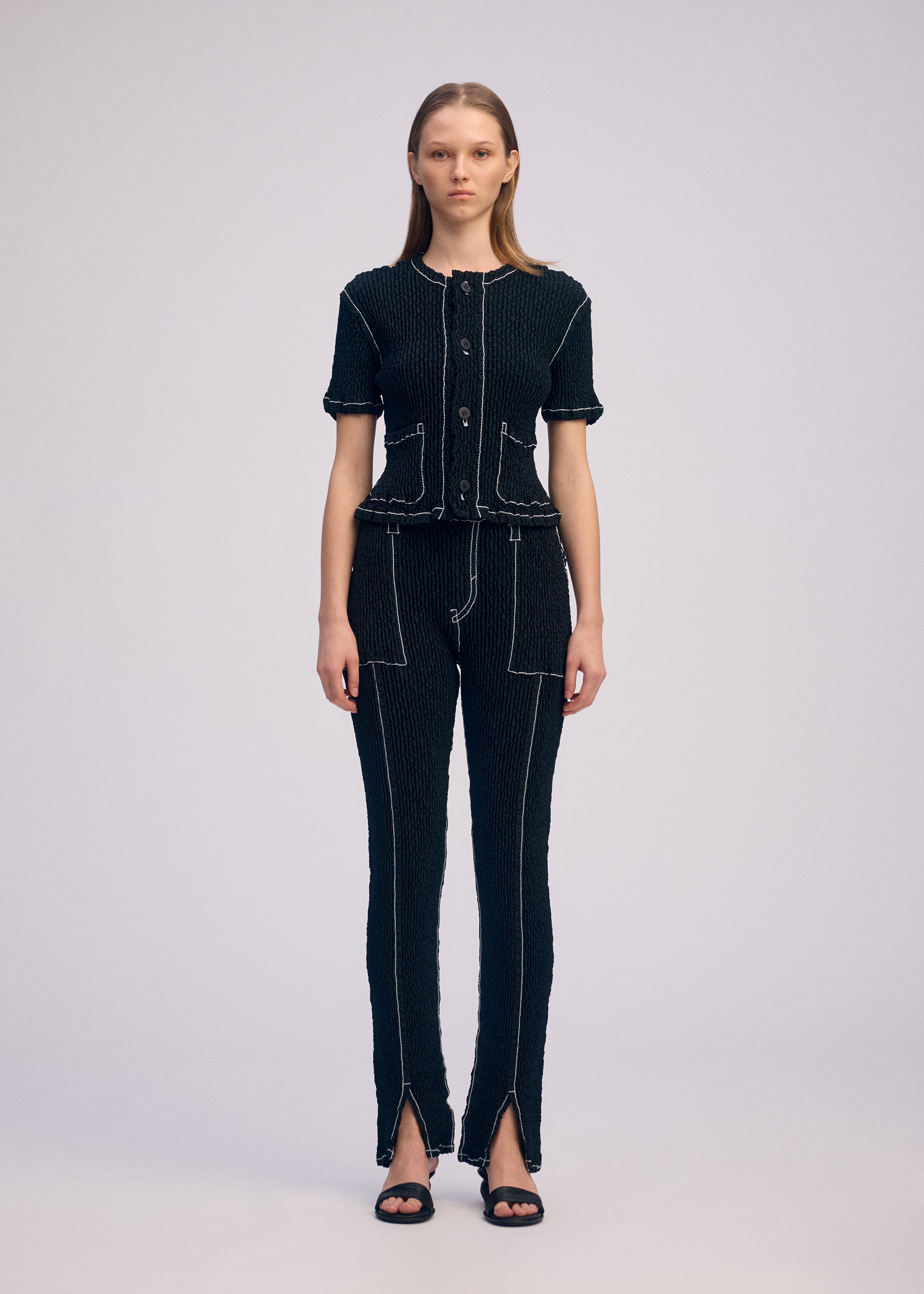 Zoomed view A woman with straight hair wears the ISSEY MIYAKE WAVELET ENSEMBLE—a jacquard stretch short-sleeved button-up top and matching pants with white stitching, front pockets, pant hem slits—and black sandals.