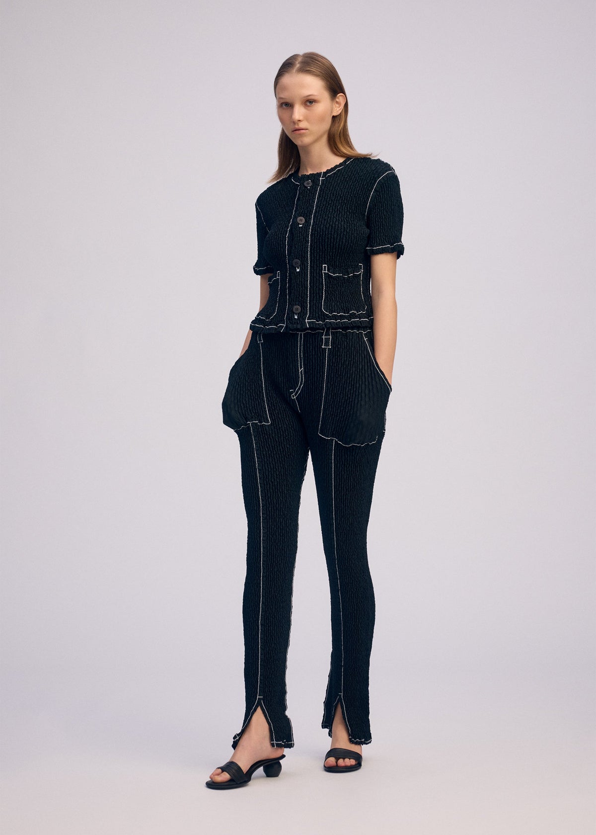 A woman stands against a plain background wearing the ISSEY MIYAKE WAVELET ENSEMBLE, a black jacquard textured short-sleeve top and pants with white stitching and front pockets, paired with open-toe black sandals. Her hands are in her pockets as she gazes sideways. view 6 of 7