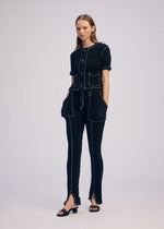 A woman stands against a plain background wearing the ISSEY MIYAKE WAVELET ENSEMBLE, a black jacquard textured short-sleeve top and pants with white stitching and front pockets, paired with open-toe black sandals. Her hands are in her pockets as she gazes sideways.