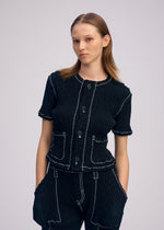 A woman with straight, shoulder-length hair stands against a plain background, wearing the ISSEY MIYAKE WAVELET ENSEMBLE—a black jacquard stretch short-sleeved cardigan and matching pants with white stitching, hands in her pockets.
