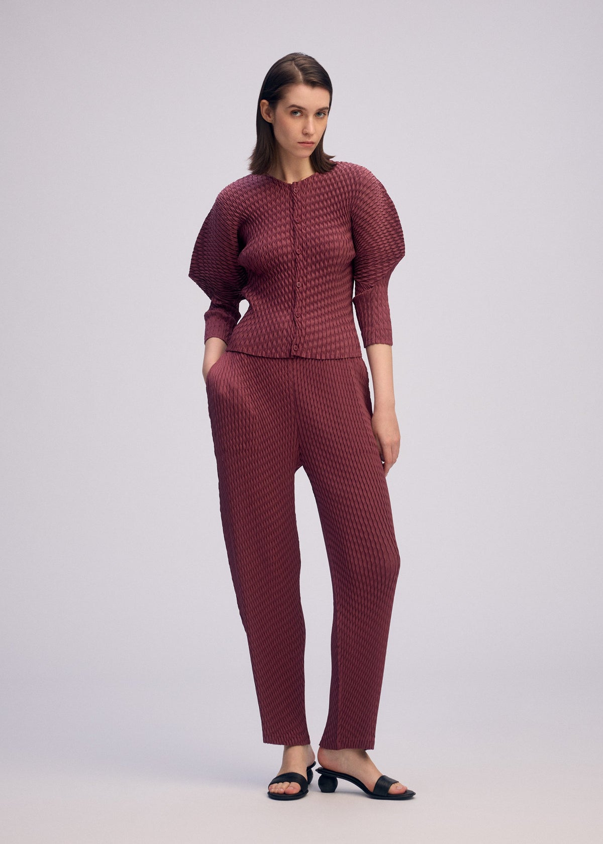 A woman poses against a plain background in the ISSEY MIYAKE SLEEK PLEATS set—wine-colored pleated cardigan and matching pants with short sleeves—styled with black heeled sandals. Her hands are in her pockets as she looks slightly to the side. view 6 of 7