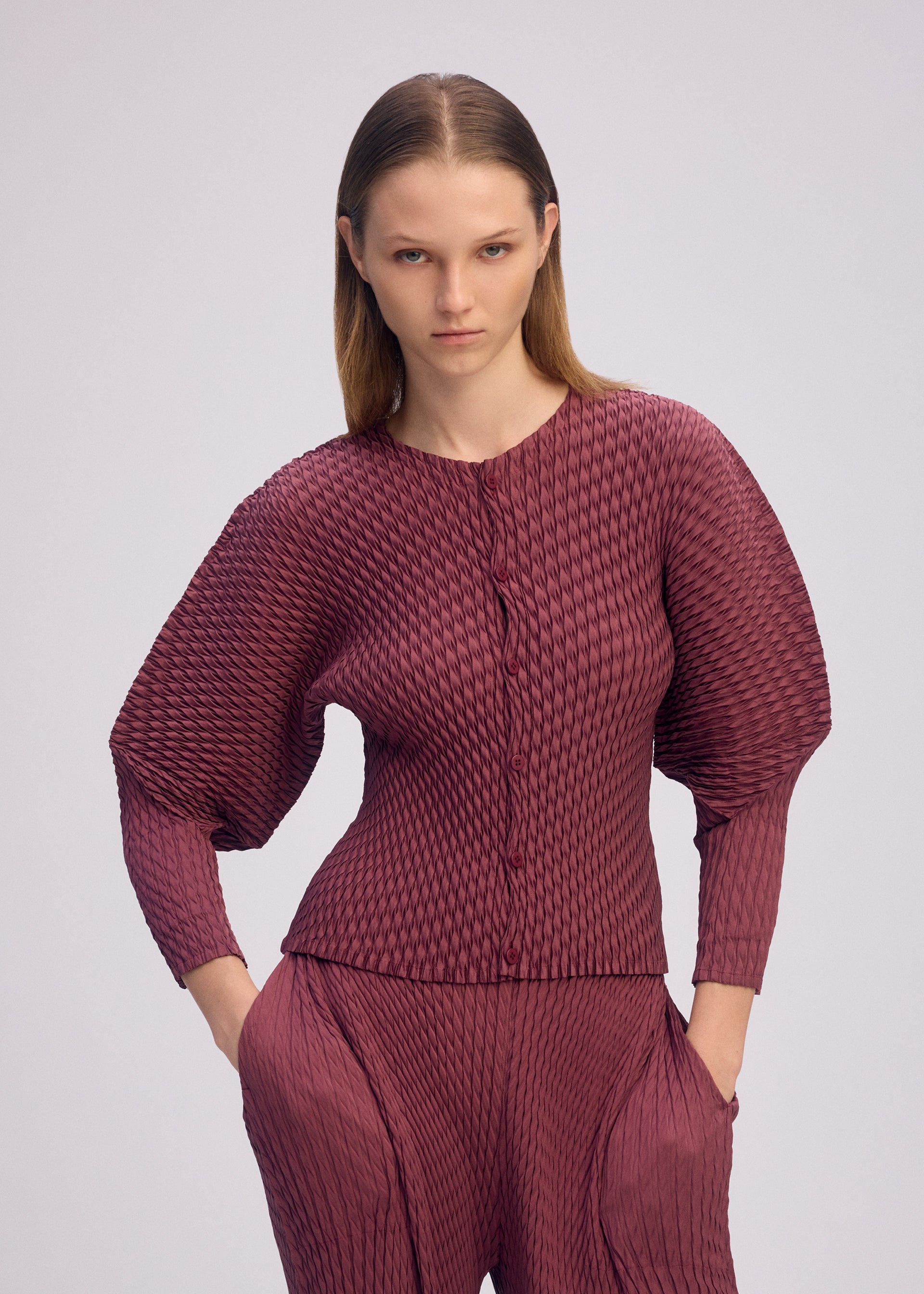 Zoomed view A woman with straight, light brown hair wears the glossy plum SLEEK PLEATS long-sleeve button-up top and matching pants by ISSEY MIYAKE, standing with her hands in her pockets against a plain light background.