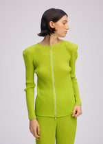A person with short brown hair wears the ISSEY MIYAKE BOX SHOULDER PLEATS—a bright lime green, long-sleeved zip-up cardigan and matching pleated pants—standing against a plain light background, looking to the side.