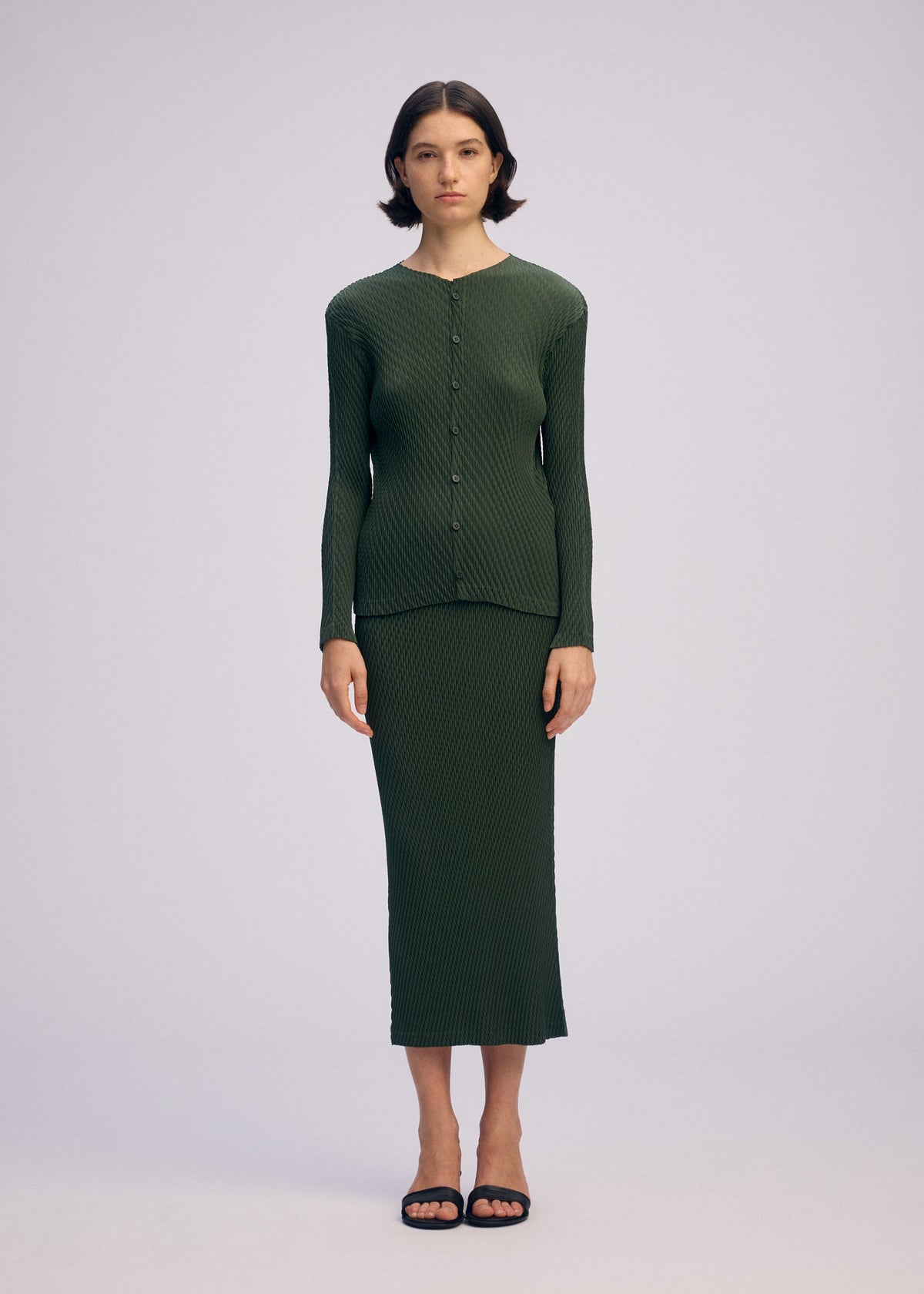 A person stands facing forward, wearing the ISSEY MIYAKE RHOMBUS PLEATS ribbed, long-sleeve, button-up dark green top and matching ankle-length skirt with black sandals against a plain light background. view 3 of 4