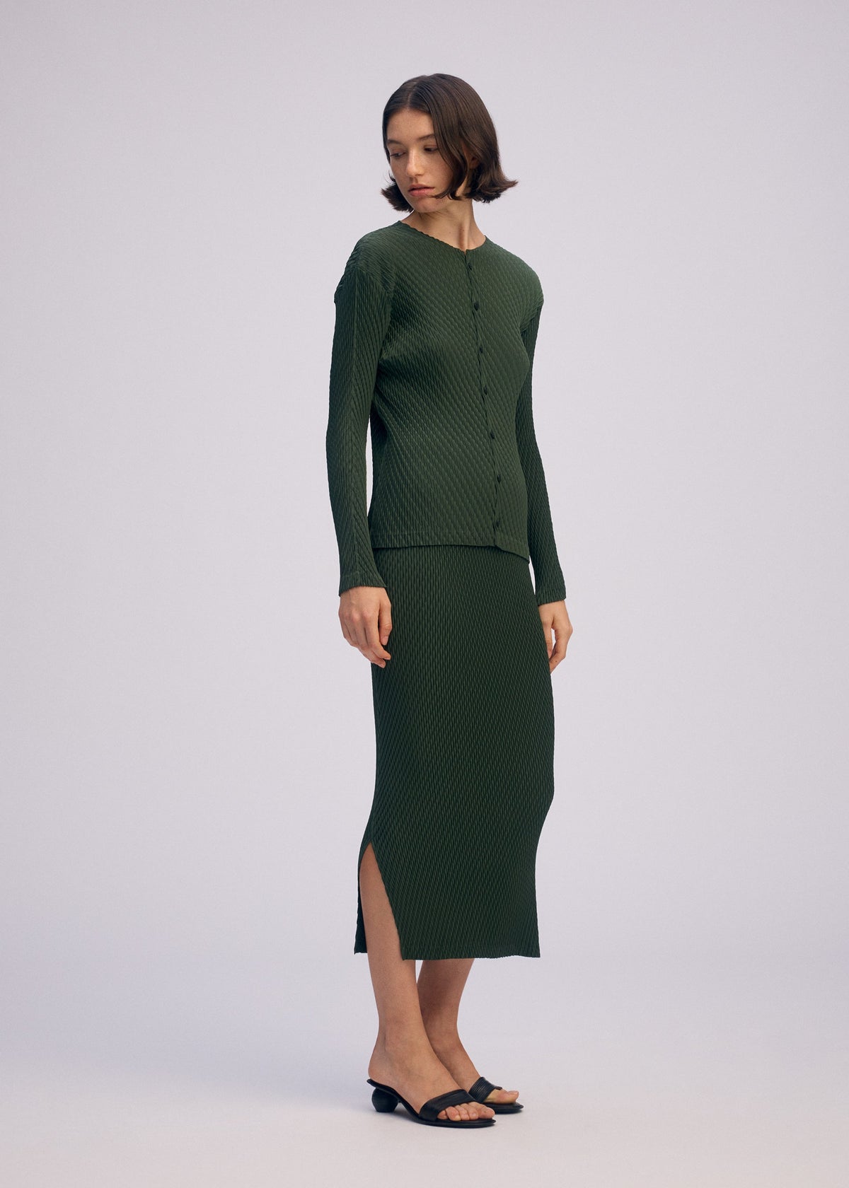 A woman with short brown hair wears ISSEY MIYAKE’s RHOMBUS PLEATS: a matching dark green ribbed knit cardigan and midi skirt with side slits, featuring a relaxed fit and sleek silhouette. She pairs the look with black heeled sandals. view 4 of 4