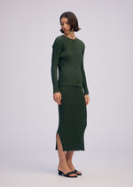 A woman with short brown hair wears ISSEY MIYAKE’s RHOMBUS PLEATS: a matching dark green ribbed knit cardigan and midi skirt with side slits, featuring a relaxed fit and sleek silhouette. She pairs the look with black heeled sandals.