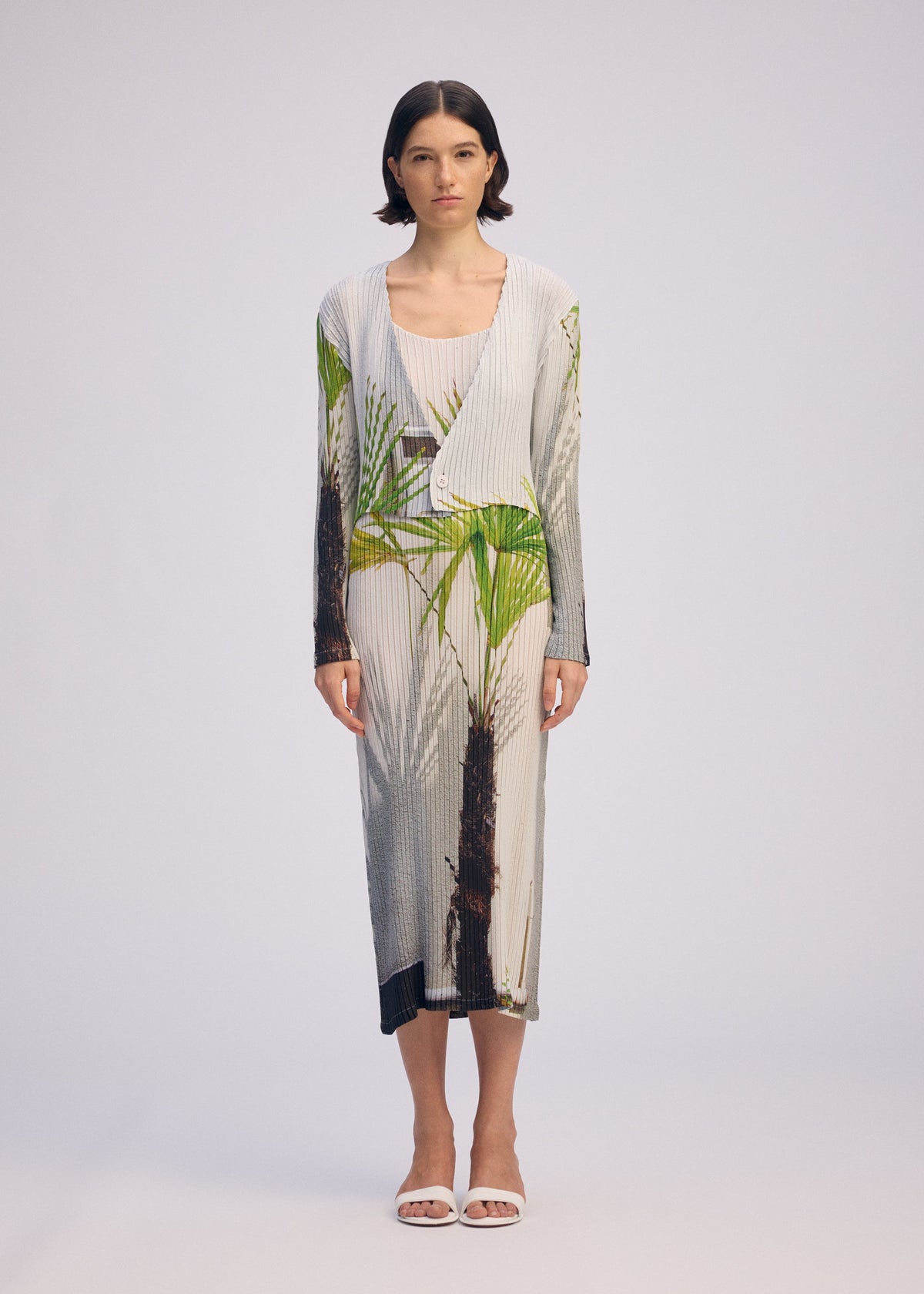 A woman with short dark hair wears the ISSEY MIYAKE URBAN JUNGLE PLEATS: a light ribbed dress and a matching cardigan with green and brown palm prints. She pairs the set with white sandals, standing against a plain light background. view 4 of 6
