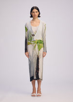 A woman with short dark hair wears the ISSEY MIYAKE URBAN JUNGLE PLEATS: a light ribbed dress and a matching cardigan with green and brown palm prints. She pairs the set with white sandals, standing against a plain light background.