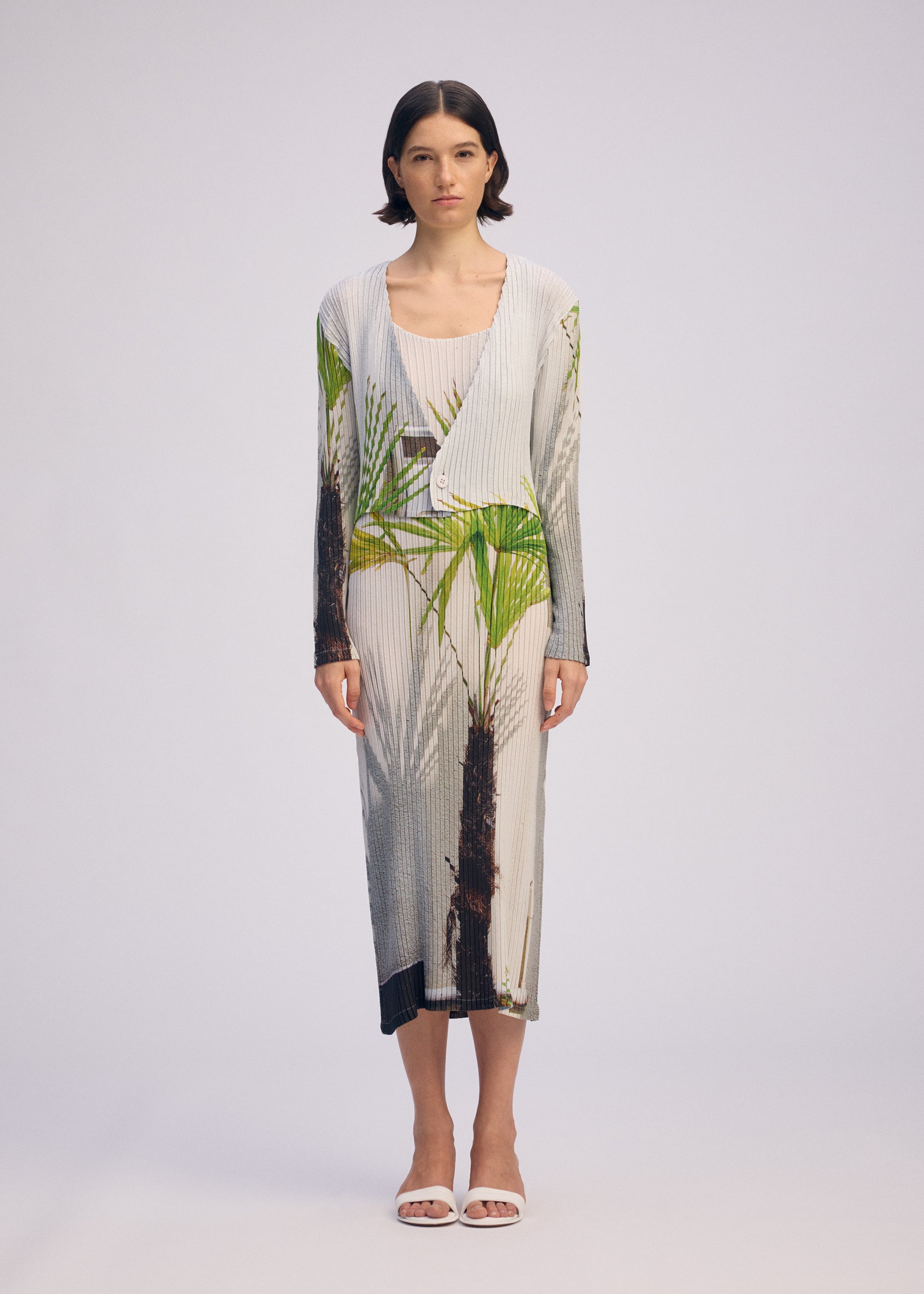 Zoomed view A woman with short dark hair wears the ISSEY MIYAKE URBAN JUNGLE PLEATS: a light ribbed dress and a matching cardigan with green and brown palm prints. She pairs the set with white sandals, standing against a plain light background.