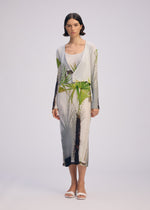 A woman faces forward in ISSEY MIYAKE's URBAN JUNGLE PLEATS, a long light pleated dress with green palm prints, layered with a matching palm print cardigan and white sandals, set against a plain light background.