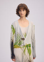 A woman with short, curly brown hair models ISSEY MIYAKE's URBAN JUNGLE PLEATS—a ribbed, light-hued cardigan and matching dress with green palm leaf prints—against a plain light background.