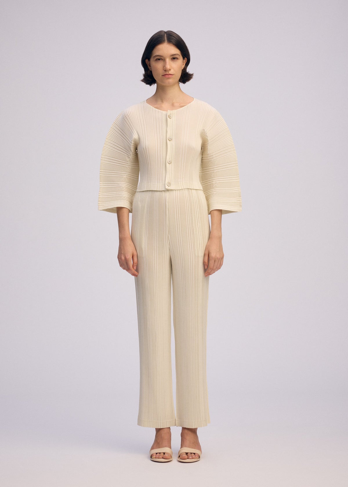 A woman with short dark hair wears ISSEY MIYAKE’s LINEN LIKE RIB PLEATS: a cream pleated cardigan with box pleats, wide sleeves, large buttons, and matching straight pants. She pairs them with open-toed sandals against a plain background. view 5 of 7