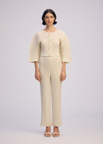 A woman with short dark hair wears ISSEY MIYAKE’s LINEN LIKE RIB PLEATS: a cream pleated cardigan with box pleats, wide sleeves, large buttons, and matching straight pants. She pairs them with open-toed sandals against a plain background.