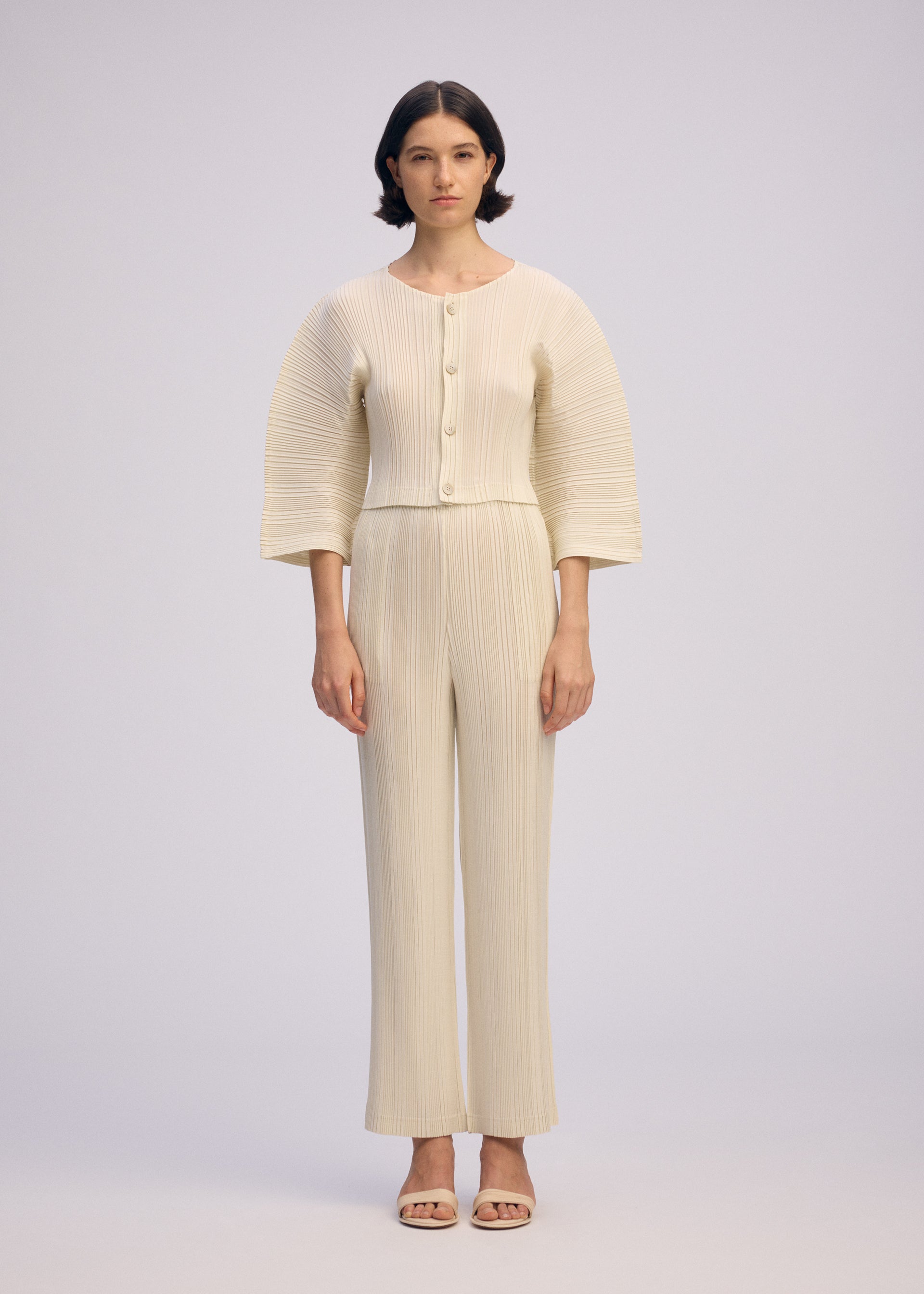 Zoomed view A woman with short dark hair wears ISSEY MIYAKE’s LINEN LIKE RIB PLEATS: a cream pleated cardigan with box pleats, wide sleeves, large buttons, and matching straight pants. She pairs them with open-toed sandals against a plain background.