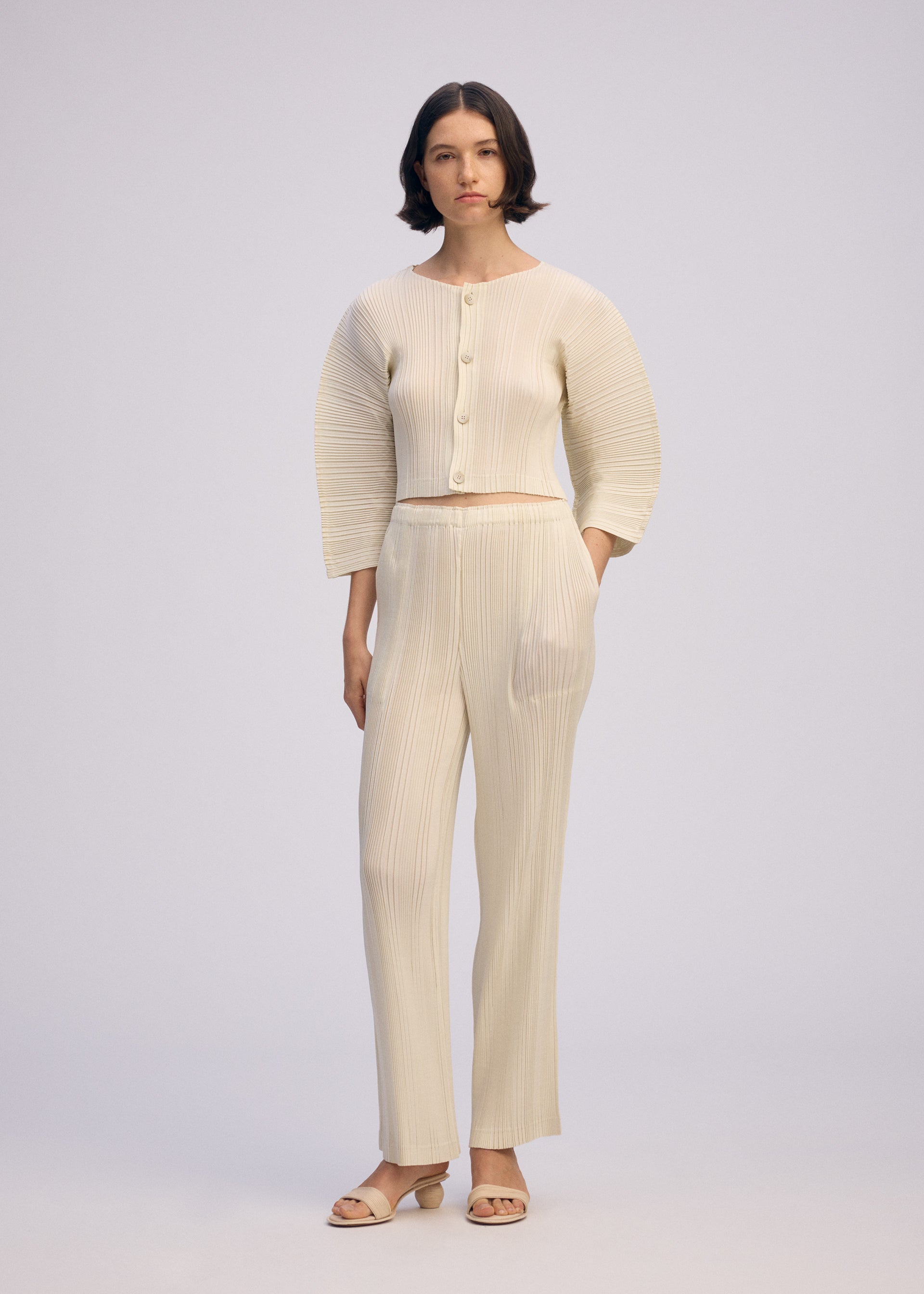 Zoomed view A woman with short dark hair and a neutral expression wears the ISSEY MIYAKE LINEN LIKE RIB PLEATS pantsuit, featuring wide sleeves, large buttons, and a linen-like texture, paired with matching heeled sandals against a plain background.