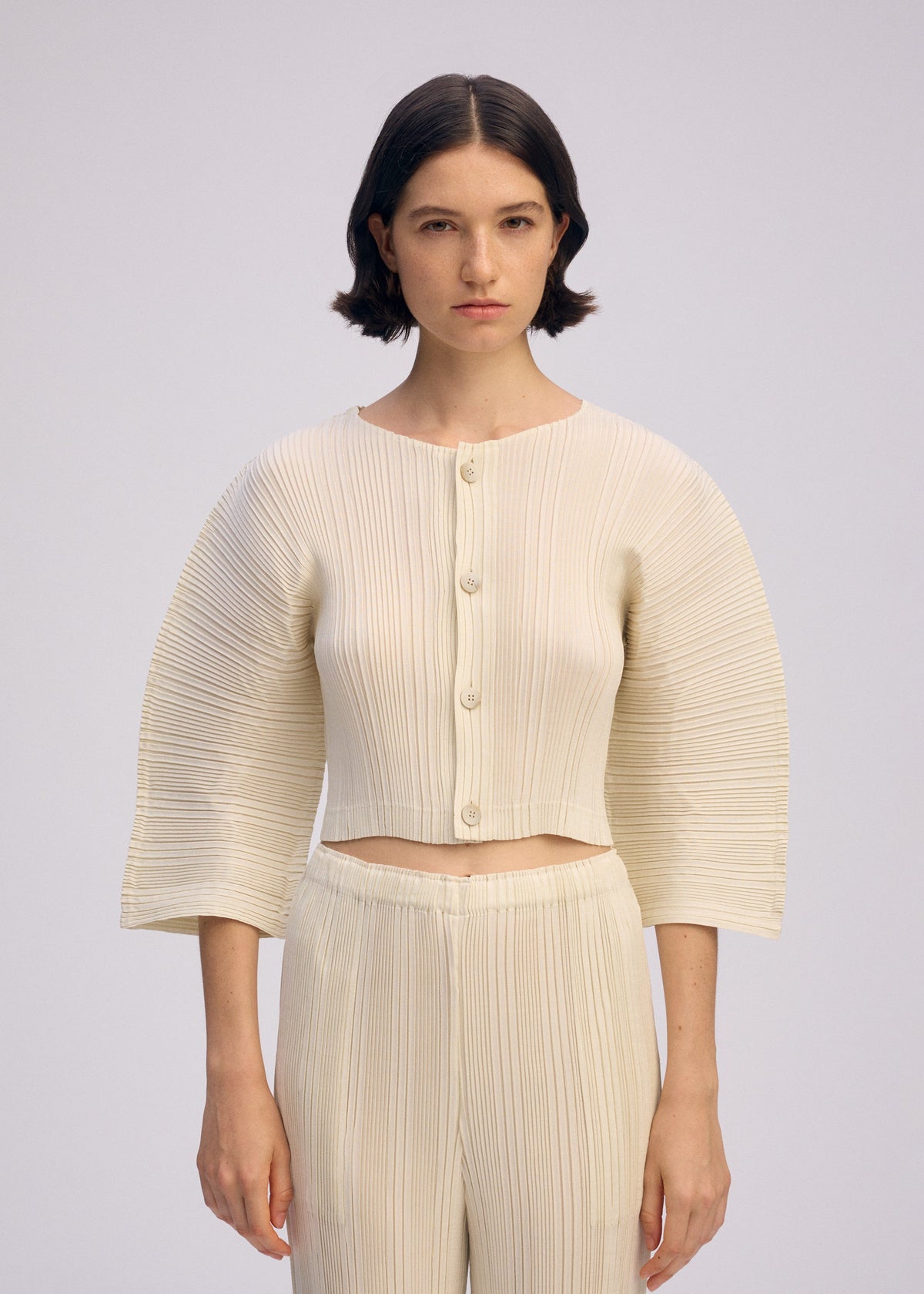A woman with short dark hair wears ISSEY MIYAKE's LINEN LIKE RIB PLEATS set—a cream box-pleat button-up top with wide sleeves and matching pleated pants—against a plain light background. view 7 of 7