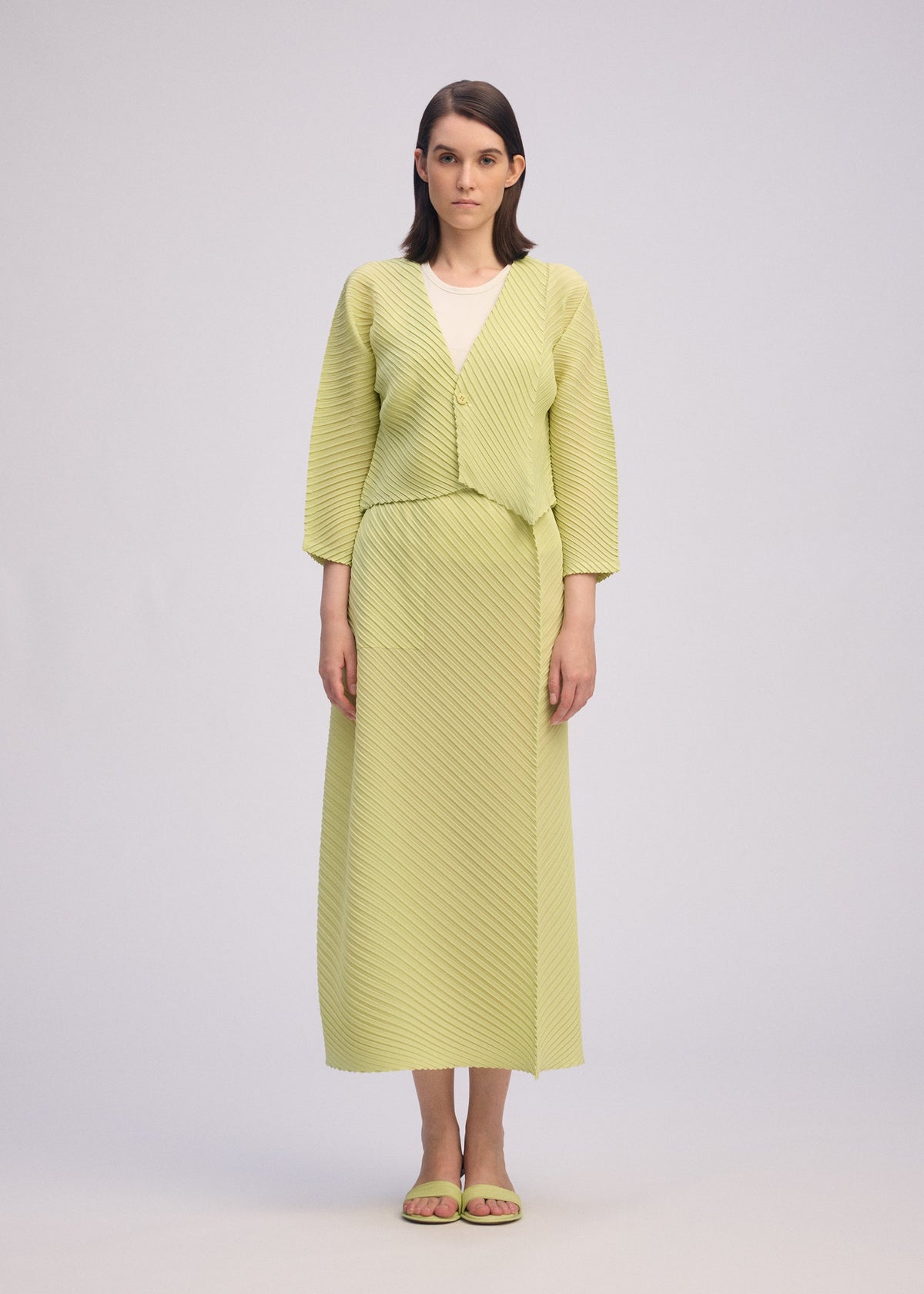 A woman faces forward against a plain background, wearing the ISSEY MIYAKE PLASTER PLEATS SOLID light green recycled polyester cardigan and matching long skirt over a white top, paired with green sandals. view 5 of 7