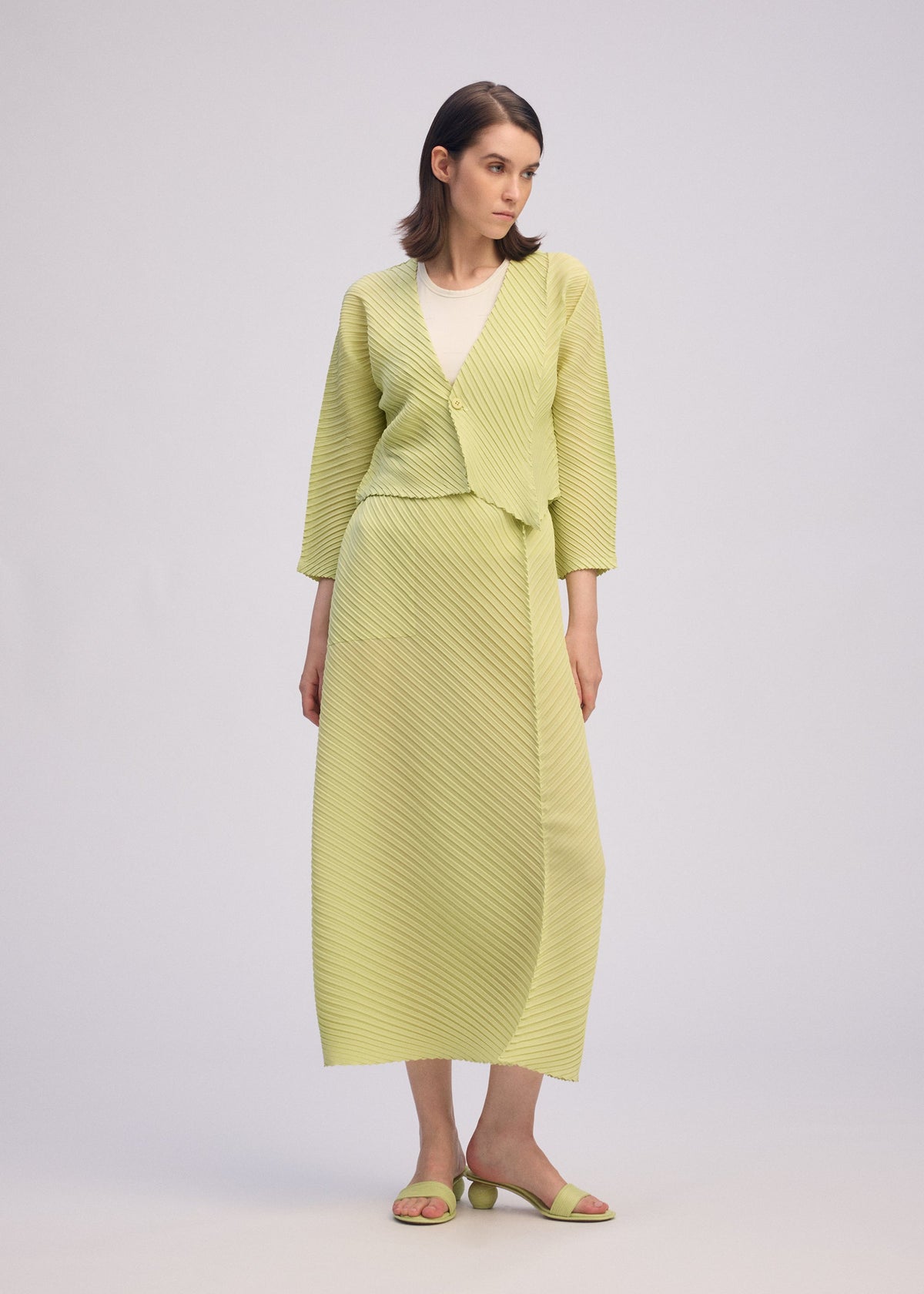 A woman models the ISSEY MIYAKE PLASTER PLEATS SOLID in light yellow-green—a crêpe de Chine long skirt with matching draped cardigan, white top, and open-toe sandals—standing against a plain background, looking to her left. view 6 of 7