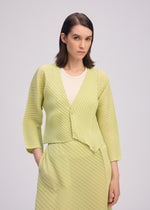 A woman with straight brown hair wears the ISSEY MIYAKE PLASTER PLEATS SOLID, a light yellow pleated crêpe de Chine cardigan, over a cream top and matching skirt, standing with one hand in her pocket against a light background.