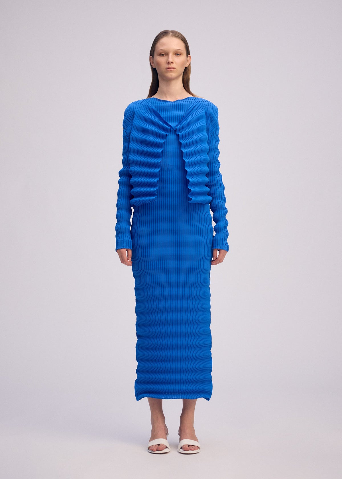 A woman faces forward in the ISSEY MIYAKE SPRING PLEATS dress—a vibrant blue, long-sleeved, floor-length piece with handmade pleats and a ribbed texture—paired with white open-toe heels against a plain light background. view 4 of 6